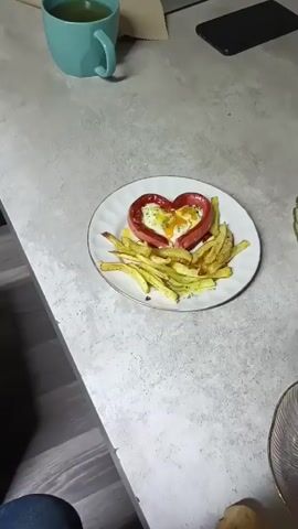 The video shows a close-up, overhead view of a white plate with a floral rim, placed on a speckled gray countertop. The plate contains a heart-shaped breakfast dish. The heart is formed by a thick, reddish sausage, with an egg cooked inside it. The egg appears to be sunny-side up, with a visible yolk. To the left of the heart-shaped dish, there is a pile of french fries. The fries are cut into thick, irregular shapes and have a golden-brown appearance, suggesting they have been fried. Some green herbs or seasoning are visible sprinkled on the fries. In the background, to the left, is a turquoise mug containing a light-colored liquid, likely tea or coffee, and a brown paper object. To the right of the mug, a black smartphone is partially visible. The lighting suggests an indoor setting.
