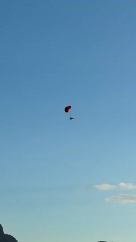 A powered parachute is seen flying in a clear blue sky. The parachute is red and the person piloting it is small and dark. There are a few white clouds at the bottom of the frame. The powered parachute is drifting slowly across the sky.