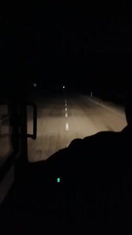 The video shows a dark road at night, illuminated by a vehicle's headlights. The road has white dashed lines down the center, and a single white post is visible on the right side.  The view is from inside a vehicle, with the door handle and the silhouette of the driver visible on the left and right sides of the frame respectively. The vehicle appears to be in motion.