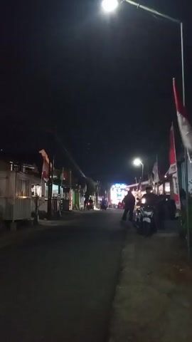 The video shows a street at night, lit by a streetlamp and a bright, possibly moving, light source in the distance. Several Indonesian flags are displayed along the sides of the street, some of which are lit from behind, creating a glow. Silhouetted figures of people and motorcycles are visible, and there appears to be a gathering or event taking place. The road surface is wet, reflecting the lights.