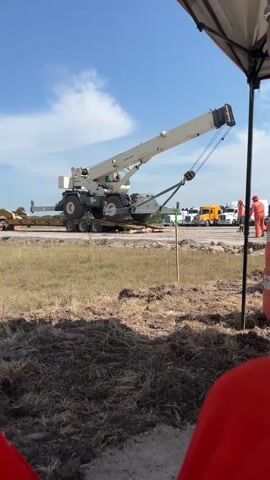 A large white crane is being transported on a flatbed trailer in an outdoor setting. The crane's boom is extended and angled upwards. A person wearing an orange uniform is visible standing near a yellow truck in the background. The ground is covered with dry grass and dirt. The sky is blue with scattered clouds. A portion of a white canopy or tent is in the foreground on the right side of the frame. The scene appears to be a construction or industrial site.