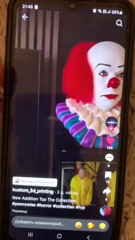 The video shows a close-up of a cell phone screen displaying a social media application. The main content of the screen is a video featuring two busts of Pennywise the clown from the movie "It." The bust on the left is of Pennywise in his classic colorful costume with red hair. The bust on the right is of Pennywise in his more menacing, blood-stained appearance. The video also shows a smaller preview window at the bottom of the screen, displaying a scene with two people wearing bright yellow jumpsuits. The user interface elements of the social media app are visible, including a username "kustom_3d_printing," hashtags like "#pennywise," "#horror," and "#collection," and engagement icons such as likes and comments. The overall context suggests a showcase of custom 3D-printed fan art.