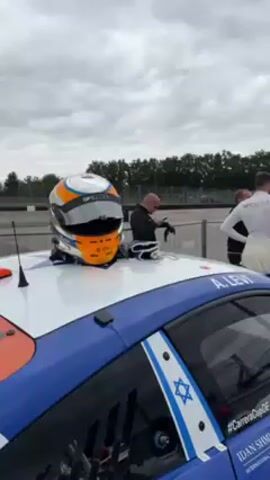The video shows a race car with a helmet and gloves on its roof. The car is white with blue and orange accents, and has the Israeli flag on the side. There is a man standing next to the car, looking at his phone. Other people are nearby. The environment is a racetrack on an overcast day.