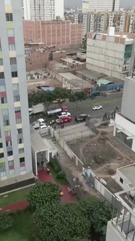 This aerial video shows a traffic incident on a multi-lane road in Breña, Peru. The scene is set during daylight hours with a cloudy or overcast sky, suggesting a mild, possibly humid climate. The urban environment is characterized by multi-story buildings, some with exposed brickwork, and a mix of residential and commercial structures.

The central focus of the video is a collision involving a red tow truck, a white bus, and a white pickup truck. The tow truck appears to be actively involved, with its equipment extended and positioned around the bus. Several individuals, some wearing high-visibility yellow vests, are gathered around the accident site, suggesting they are emergency responders or involved parties assessing the situation. Two individuals in what appear to be dark uniforms are also present. The damaged vehicles are positioned on the road, indicating a disruption to traffic.

In the background, other vehicles, including a red and a white truck, as well as yellow taxis, are visible on the road, some appearing to be moving slowly or stopped. The presence of a tanker truck further down the road suggests a variety of commercial vehicles operate in this area. A fenced-off area with some construction materials or debris is visible in the foreground, alongside a green, leafy area with a red bench.

While no explicit emotions are conveyed due to the distance and nature of the recording, the scene depicts a busy urban street dealing with the aftermath of a vehicular accident, likely causing traffic delays and requiring immediate attention from authorities and recovery services. No legible text is visible on the vehicles or surrounding structures in this particular clip.