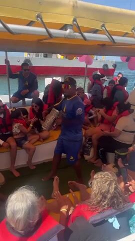 Here is a description of the video:

The video shows a lively scene onboard a boat, likely on a tour or cruise.  A group of people of various ages are visible, most wearing red life jackets.  A man in a blue shirt and shorts, wearing sunglasses and a cap, stands in the center of the group, playing a percussion instrument (appears to be a guiro).  Other men on the boat are seated and playing drums.  The people are smiling and enjoying the music.  The boat has a yellow roof and is surrounded by water.  Another boat with a red and white hull is visible in the background.  Balloons decorate the background.