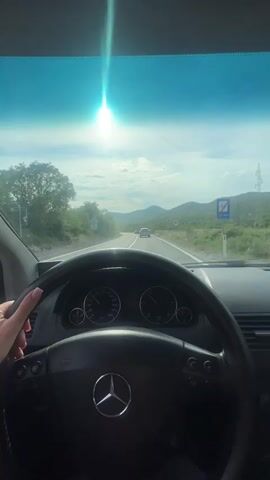 A person is driving a Mercedes Benz on a highway, presumably during the day. The road is straight, with green hills and vegetation on either side. There's another car a distance ahead. The sun is high in the sky, with glare on the windshield. A hand is visible on the steering wheel, nails neatly manicured. The dashboard, steering wheel, and the Mercedes-Benz logo are clearly visible. There is a speed limit sign visible on the right side of the road.