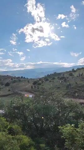 This media file captures a lively outdoor scene, likely a festival or event, set in Antarut, Armenia. The camera pans across a grassy, hilly landscape under a bright, partly cloudy sky. The sun shines through breaks in the clouds, casting rays and highlighting the terrain.

The primary subjects are groups of people scattered across the hillside, some standing, others seemingly walking or milling about. They are dressed in casual attire, suggesting a relaxed atmosphere. In the foreground, dense green foliage frames the lower part of the shot.

The scene appears to be an outdoor gathering, possibly an event like a rally, race, or cultural festival, given the presence of what looks like a marked course or boundary tape (white and orange ribbons) stretching across the grass in several places. There are also some vehicles visible in the middle ground, further suggesting an organized event. In the distance, across a valley, a historical settlement or fortress is perched on a hilltop, its silhouette adding a sense of history and grandeur to the modern gathering.

The time of day is daytime, with bright sunlight and clear skies interspersed with fluffy white clouds. The weather seems pleasant, ideal for an outdoor event. The rolling hills and distant mountains are characteristic of the Armenian landscape. There are hints of text on banners at the bottom left of the frame, one appears to be red and yellow with some lettering, but it's too blurry to decipher. The overall mood conveyed is one of community and enjoyment, with people participating in or observing an event in a beautiful natural setting.