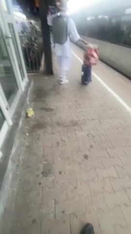 A person wearing white clothing and a hat stands on a tiled platform next to a train. They are seen from the waist down, and are holding a black and red bag in their right hand, which is attached to a rolling suitcase. They are also wearing white sneakers with black accents. The platform is made of light grey tiles, and appears to be outdoors, possibly at a train station, with a train visible on the right side of the frame. There is some debris on the ground. The video is shot from a low angle, looking up at the person.