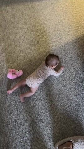 A baby in a pink floral onesie lies on a gray carpet. The baby is on their stomach, pushing up on their hands, trying to crawl. There is a pink slipper nearby.