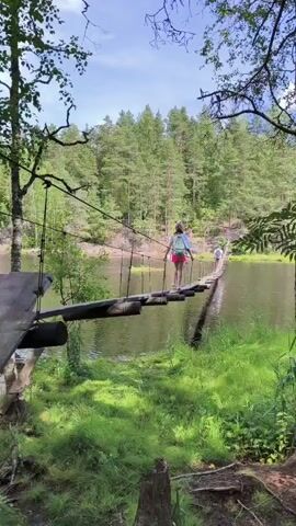 A young person wearing a pink hat, light blue top, and pink shorts, carrying a light blue backpack, walks across a long, narrow, suspended wooden plank bridge over a body of water. The bridge is supported by ropes and poles and appears to be rustic. Another person is visible further ahead on the bridge. The surroundings are lush and green, with tall trees and vegetation on the banks of the water and a dense forest covering the opposite shore. The sky is partly cloudy. The video is shot from a low angle, focusing on the person crossing the bridge.