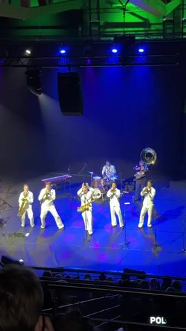 A band of seven musicians are performing on a dimly lit stage with blue and green spotlights. The band members are all wearing matching white outfits. Six of them are standing at the front of the stage, playing various brass instruments, including trumpets and saxophones. One musician in the center is playing a saxophone and appears to be singing. Behind them, a drummer is playing a drum set, and another musician is playing a tuba. The stage floor has circular light patterns projected onto it. In the foreground, the audience is visible, with some people raising their hands. The background is a dark blue curtain.