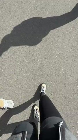 A person is walking on an asphalt road, with a view of mountains in the distance. The person is wearing black pants, and a shoe in a beige color with white laces.  The shot begins with the person’s shadow on the road, and then shows the person walking along the road, in the direction of the mountains. There are trees along both sides of the road. Other people can be seen in the distance, also walking along the road. There is a white vehicle parked off the side of the road. The mountains are covered in snow, and the sky is cloudy.