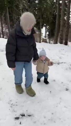 A woman wearing a black puffer jacket, light blue jeans, and sage green shoes walks in the snow with a young child. The child wears a light blue knit hat, a tan puffer vest, dark blue pants, and black boots. The woman appears to be holding the child’s hand to help them walk on the snow-covered ground. Trees are visible in the background. The woman also wears a light gray hat or hood.