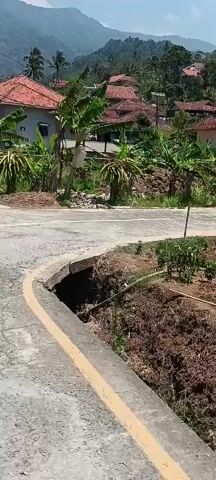 The video shows a road with a significant portion of its edge missing, creating a deep drop-off. The road appears to be paved and has yellow and white lines painted on it. The surrounding environment is rural, with houses and lush vegetation, including banana trees, visible in the background. A car is seen driving on the road in the distance. The ground near the collapsed edge of the road is bare earth and some scattered debris. The day appears to be sunny. The video is shot from a moving perspective, suggesting someone is walking or driving along the road and filming.