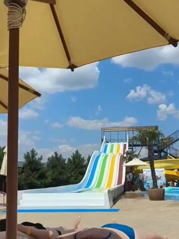 The video shows a water park on a sunny day. There are several colorful water slides, a swimming pool, and palm trees. People are seen in and around the water slides and pool, enjoying the attractions. Umbrellas provide shade for the lounging areas. The sky is blue with scattered clouds. The video appears to be shot from a seated perspective, possibly from someone relaxing on a lounge chair.