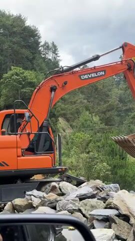 The video captures a scenic mountainous landscape. Initially, a large orange excavator is visible, partially obscured by a vehicle's side mirror. The camera then pans across a valley, revealing terraced rice paddies descending a hillside, with a small stream flowing at the bottom. The surrounding area is densely covered in green trees and vegetation, indicating a lush, rural environment. The video concludes with a close-up shot of a dirt path and more greenery.