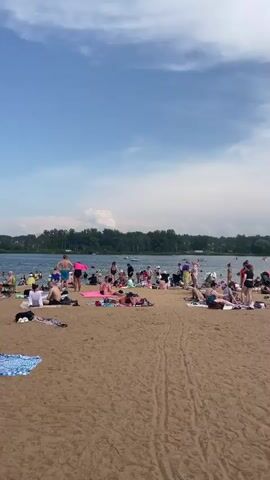 The video shows a crowded beach on a sunny day. The sand is a light brown color and there are many people sunbathing, walking, and interacting on the beach. Some people are in swimwear, while others are wearing casual clothing. There are towels and blankets spread out on the sand. In the background, there is a body of water, likely a lake or ocean, with more people visible in the water and on boats. Lush green trees line the far side of the water. The sky is blue with scattered white clouds. The overall atmosphere appears to be relaxed and recreational.