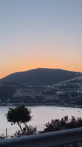 The video captures a scenic coastal landscape during sunset, likely filmed from a moving vehicle. In the foreground, there are green trees and shrubs, along with a dark road barrier or railing. A light pole is visible on the right side of the frame.

The mid-ground features a calm body of water, possibly the Mediterranean Sea, reflecting the soft light. Several small boats are scattered across the water. On the hills rising from the coastline, numerous white buildings are visible, suggesting a town or resort area.

In the background, a large mountain or hill dominates the horizon, silhouetted against the setting sun. The sky exhibits a beautiful gradient, transitioning from warm orange and yellow hues directly above the mountain to a pale blue at the top. The overall ambiance suggests a peaceful evening in a picturesque location.

The location is Kaş, Turkey, and the video shows the characteristic blend of mountains, sea, and hillside architecture typical of the region. No visible text is present in the video.