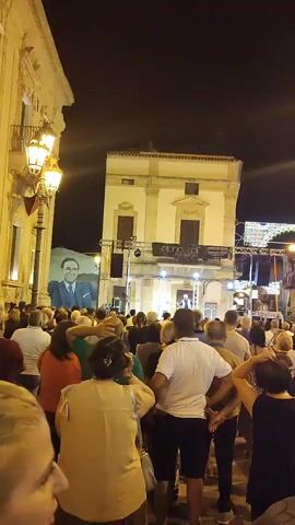 A crowd of people are gathered at night in an outdoor square. A large portrait of a man in a suit is visible on the left side of the frame, and a stage with a banner is set up in front of a building in the background. Several people are standing on the stage, and some are speaking into microphones. The scene suggests a public event or performance.