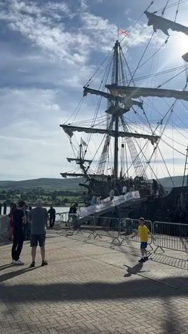 A tall ship with multiple masts and sails is docked at a pier under a partly cloudy sky. Several people are visible on the deck of the ship and on the pier. A boy in a yellow shirt and blue shorts runs in the foreground. Metal barriers line the edge of the pier. In the background, rolling hills are visible. The sun shines brightly, casting shadows.