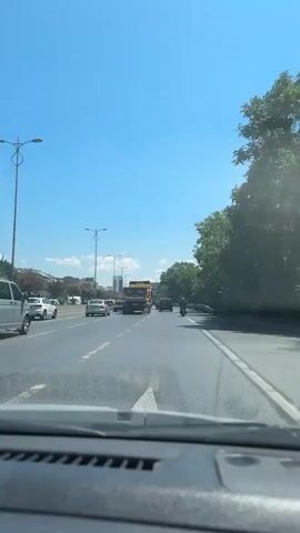The video is filmed from the dashboard of a vehicle moving on a multi-lane road under a clear blue sky. The road appears to be a highway or a major thoroughfare. On the left side of the road, there are other vehicles, including a white van and several cars. Further in the distance, there are buildings and trees. On the right side of the road, there is a dense line of trees. In the middle of the road, several vehicles are visible, including a large yellow truck. The camera perspective is from the driver's point of view, showing the dashboard in the foreground. The scene is one of daytime traffic on a sunny day.