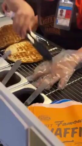 Here's a concise description of the video:

Two workers at a waffle stand prepare waffles. One person uses tongs to move a waffle from the rack to a plastic bag. They take an orange paper bag, possibly for packaging, and hand it to the other worker. There are several stainless steel containers with sauces or fillings behind the rack. The waffles are round and have a grid pattern. The workers are wearing orange and black uniforms and have name tags.