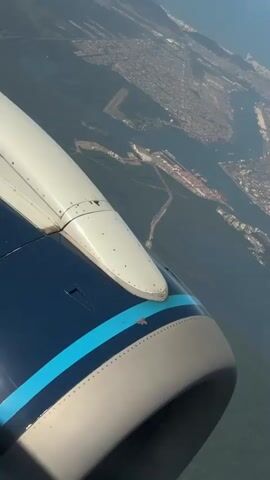 A point-of-view video captures an aerial view from an airplane window. The aircraft's blue and white engine and wing are visible in the foreground. Below, a sprawling city with numerous buildings lines a coast next to a large body of water. Mountains and hills are also visible in the distance, partially covered by scattered clouds in a clear blue sky.