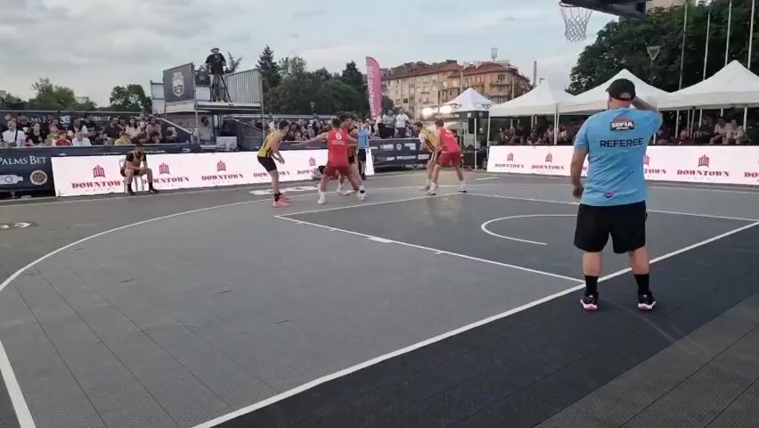 A wide shot shows a 3v3 basketball game in progress on an outdoor court. Six players, three in red jerseys and three in yellow and black jerseys, are actively playing. A referee in a blue shirt stands near the sideline, observing the game. A crowd of spectators is visible in the background, seated on bleachers and standing around the court. White tents and sponsor banners, including "PALMS BET" and "Domino's Concept," are also visible in the background. The court is black and gray, with white lines marking the boundaries and key. The weather appears overcast.