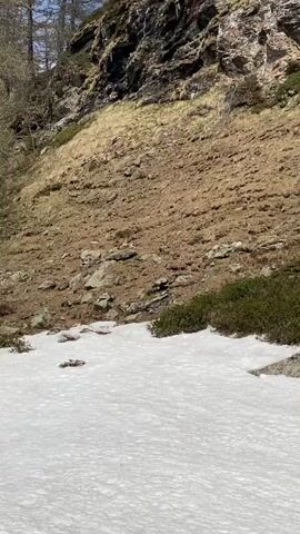 The camera is held still in a nature shot. There is a patch of snow in the foreground. Above that is a hillside covered in brown grass and dirt. At the top of the hill is a craggy rock formation.  The sky is visible at the very top of the frame, appearing blue.