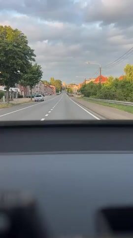 The video captures a first-person perspective from inside a vehicle, looking forward through the windshield. The road ahead is a straight, paved two-lane road with a white dashed center line and solid white lines marking the edges. On the left side of the road, there are several buildings, some appearing to be houses or small businesses, with trees lining the sidewalk. On the right side, there are more buildings, greenery, and a metal guardrail. The sky is partly cloudy with a mix of grey and white clouds, and the lighting suggests it is either early morning or late afternoon. There are a few cars visible on the road and parked along the side. The overall impression is that the vehicle is driving through a moderately populated area, possibly a town or village, on a pleasant day.