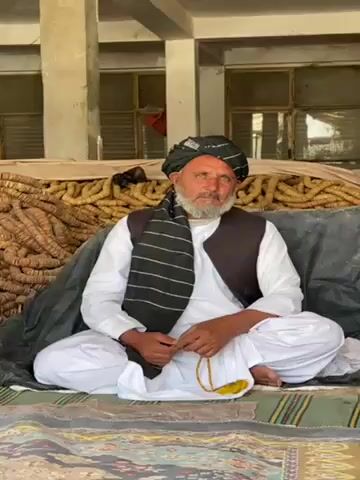 An eye-level shot features an older man with a grey beard and mustache, seated cross-legged on a patterned rug. He wears a white shalwar kameez, a dark vest, and a black turban with white stripes. In his hands, he holds a string of yellow prayer beads. The man is looking off to his right.

Behind him is a pile of what appears to be dried organic material stacked against a wall. The setting appears to be an open-air structure with concrete pillars and a window with a metal frame visible in the background. Another person’s speech can be heard in the video.