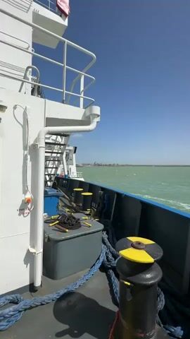 The video captures a view from the deck of a ship on a sunny day. The camera is positioned on the ship's side, looking out over the water. The deck features white railings, a set of stairs, and several pieces of equipment including a grey storage box, blue ropes coiled on the deck, and a black and yellow bollard. A red button labeled "STOP ENGINE" is visible on the ship's structure.

The water is a greenish-blue, with visible ripples and small waves, suggesting movement. In the distance, a faint coastline or port infrastructure can be seen. A small boat, possibly a patrol or pilot boat, passes by the ship, moving from left to right across the frame. The sky is clear and blue. The sound of wind or the ship's movement is audible throughout the video.