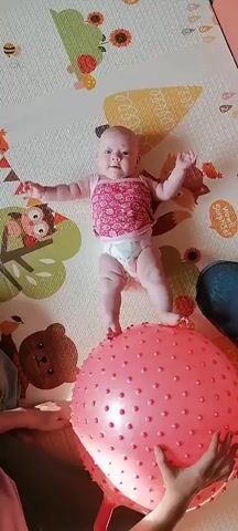 A baby is standing on a large pink exercise ball. The baby is wearing a pink top with a strawberry pattern and a diaper. The baby is looking at the camera with its arms outstretched. The baby is on a mat with cartoon animals on it. There are two hands holding the ball from below, suggesting someone is assisting the baby. The camera is positioned above the baby, looking down. The lighting is bright and natural.