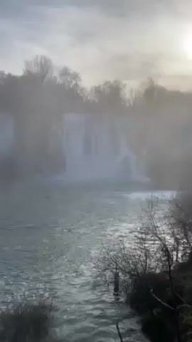 The video shows a waterfall cascading into a river, captured under a cloudy sky. The scene appears somewhat hazy, possibly due to mist or spray from the waterfall. Trees are visible along the upper bank of the river, with bare branches suggesting it may be winter or early spring. The river's surface is turbulent, reflecting the daylight above. In the foreground, there's a sparse bush, and the camera slightly shifts its position, revealing more of the scenery.