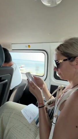 A woman with blonde hair, wearing sunglasses and a white tank top, sits in the back seat of a vehicle, holding a white smartphone and looking at it. She is wearing a patterned skirt. A person with dark hair is visible in the seat in front of her. Through the window, a body of water, a boat, and a coastline with trees can be seen. The interior of the vehicle is white.
