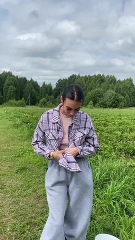The video shows a young woman standing in a field of green plants, likely strawberries, with a forest in the background and a cloudy sky overhead. She is wearing a plaid shirt over a pink top and grey sweatpants. The woman is looking down and appears to be tucking something into her pants. To the right of the woman, there is a white bucket on the ground. The field has rows of plants separated by grass, and in the distance, a line of trees marks the edge of the field.