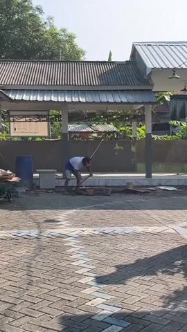In Sugih Waras, Indonesia, under a clear sky suggesting a bright, sunny day, a man is engaged in construction work. He is seen wearing a white t-shirt, blue shorts, and a red cap, his posture indicating physical exertion as he bends over a pile of materials. The scene unfolds in an outdoor setting, possibly a construction site or a renovation area, with a covered structure that has a corrugated metal roof supported by grey pillars.

In the foreground, the ground is paved with brick pavers, some of which have white markings, suggesting a designated area. To the left, a large blue barrel and a concrete container are visible. Scattered around the work area are various construction debris and tools, including what appear to be bricks and possibly cement or mortar.

The man is actively working with a tool, possibly a trowel or a hammer, in his hands, seemingly mixing or laying materials. The overall atmosphere suggests a typical daytime construction activity. While no specific emotions are overtly displayed, the man's focused posture implies dedication to his task. The backdrop features lush green foliage and the roof of another building, contributing to the rural or semi-urban Indonesian setting. There is no discernible text visible in the media.