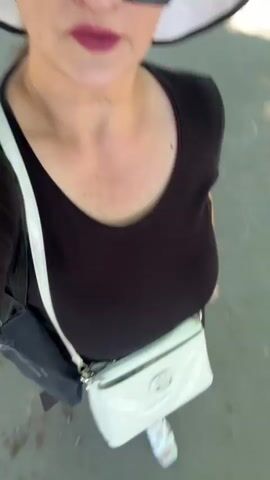 A close-up, slightly blurry video shows a woman walking outdoors on a paved path, likely during the daytime. She is wearing a black t-shirt, dark pants, a white wide-brimmed hat, and large black sunglasses. A white purse with a silver chain strap is slung across her body, and a gray bag is visible on her left shoulder. Her lips are painted with a dark pink lipstick. The background shows trees with dappled sunlight filtering through the leaves, and a building with red accents is visible in the distance. The woman's head and upper body are the primary focus, with the camera angle looking down at her as she walks.