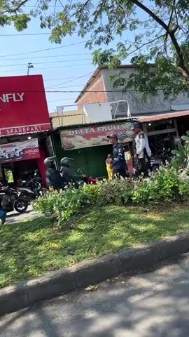 The video shows an outdoor scene during the day. In the foreground, there is a curb and a patch of grass with some low-lying plants. Beyond the greenery, several people are present. Two individuals wearing helmets are seated on what appear to be motorcycles or scooters, facing each other. One of them is holding a red object.

Further back, near a building with a red sign that says "INFLY SPAREPART," three other people are standing. Two of them are wearing uniforms that suggest they might be security personnel or employees. They appear to be in conversation with a third person who is holding a white bag. To the right of these individuals, there is a stall or shop with a green awning and a sign that reads "DELTA FRUITS." Several motorcycles are parked in front of this stall. The background shows more buildings and trees, indicating an urban or commercial setting. The lighting suggests it is a sunny day.