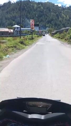 The video shows a first-person perspective of someone riding a motorcycle down a paved road. The road has some potholes and cracks. On either side of the road, there is vegetation and some buildings, including what appears to be a gas station with a large sign. In the distance, there are mountains covered in trees, and the sky is blue with some white clouds. The video is shot during the daytime.