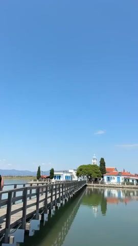 The video shows a wooden walkway extending into a body of water. On the other side of the water, there is a cluster of white buildings with red roofs, a white church with a blue dome, and several trees. The sky is clear and blue, with a few wispy clouds. The water is calm and reflects the buildings and sky. The video appears to be shot on a sunny day.