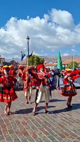 This video captures a vibrant cultural performance taking place on a cobblestone street in Cuzco, Peru. The scene is filled with people dressed in colorful traditional clothing, engaged in what appears to be a lively dance or procession. The primary subjects are individuals adorned in bright red hats and intricately patterned ponchos and skirts, many featuring geometric designs and tassels in shades of red, white, yellow, and black. Their attire is festive and elaborate, suggesting a special occasion.

The activity is clearly a performance, with dancers moving in unison, their arms extended and bodies in dynamic poses. The central figure, wearing a distinctive red, wide-brimmed hat and a richly decorated poncho, seems to be leading the group. In the background, other performers can be seen, some holding what appear to be musical instruments like drums, and others carrying staffs or poles adorned with rainbow-colored flags. Spectators line the street, observing the performance, some with smiles on their faces, indicating enjoyment and a festive atmosphere.

The setting is an urban environment with colonial-era architecture visible in the background, characterized by terracotta roofs and whitewashed walls. Mountains are visible in the distance, under a bright blue sky dotted with white, fluffy clouds. The presence of rainbow flags, often associated with LGBTQ+ pride and also with indigenous cultures of the Andes, adds another layer of cultural significance. A tall lamppost stands prominently in the middle ground, and further back, there's a green flag and a white barrier, possibly marking the performance area or a viewing section for attendees. The time of day is clearly daytime, likely late morning or afternoon, given the strong sunlight casting shadows on the cobblestones and illuminating the vibrant colors of the costumes. The weather appears to be pleasant and sunny, contributing to the overall cheerful mood of the event. There is no visible text in the frame.