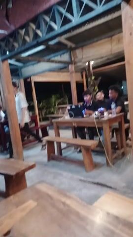 The video captures an evening scene inside a rustic, open-air cafe or eatery in Geulumpangmeujim-Jim, Indonesia. The establishment features a simple yet robust structure, characterized by wooden beams, a corrugated metal roof, and a visible section of red brick. The flooring appears to be concrete.

Several young men are present, enjoying the casual atmosphere. In the foreground, a group of three men are seated around a wooden table. One man is actively using a laptop, while the others observe and engage in conversation, with one man gesturing. A water bottle and a pair of headphones (with a visible "Monster" logo) are on the table beside the laptop. The number "12" is faintly visible on the side of this table.

Further back, another man is seen seated at a separate wooden table, also interacting with a laptop. In the background, two more men are visible sitting at another table. The overall activity suggests a relaxed gathering, possibly for study, work, or socializing. The time of day is clearly night, indicated by the artificial lighting illuminating the interior and the darkness beyond the open walls, where some distant trees and foliage can be seen. The people appear calm and engaged in their respective activities.