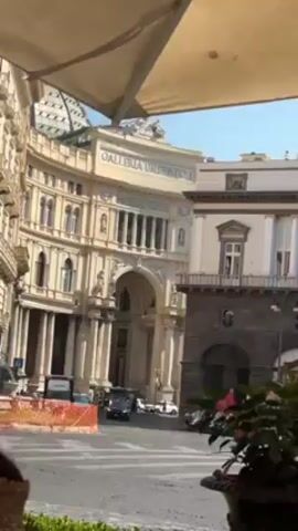 From under a canvas awning, a view of the Galleria Umberto I in Naples, Italy is visible on a sunny day. The building is constructed in a neoclassical style, with columns and arched entrances. A man crosses the street in front of the building, and there are several cars and a scooter driving nearby. The ground is paved with cobblestones. There is a potted plant with pink flowers visible in the foreground.