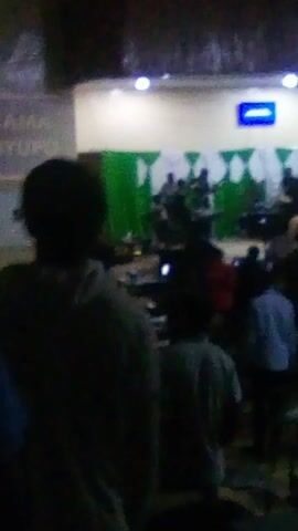 The video shows a crowded room with many people sitting on the floor. In the foreground, an individual's shoulder and back of their head are visible, obscuring some of the scene. A wall on the left side of the frame is decorated with a pattern of green and white shapes, and a blue light with the number "1" is mounted on the wall. The people in the room are mostly dark silhouettes, though some are wearing lighter colored shirts or jackets. Many appear to be looking at something out of frame. The lighting is dim, and the video is slightly blurry.