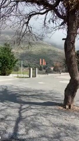 The video shows a static shot of a landscape in Italy, taken in daylight. The camera pans from behind a tree with bare branches, with a mountainous backdrop obscured by clouds in the distance. It moves to reveal a small stone church with a bell tower, surrounded by a black iron fence. There is a smaller stone building to the right of the church, also enclosed by the same type of fencing. The ground is covered with small gray pebbles. The sky is partly cloudy, with patches of blue visible.