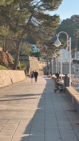The scene appears to be outdoors, likely a seaside promenade. A tall wall runs along the right side of the frame, casting a long shadow. Above the wall, a row of lamp posts with curved arms and white shades are visible. People are present on the promenade, including one person in a wheelchair. On the left, there is a stone path and a tree with green foliage. Buildings are visible in the background beyond the tree. The lighting suggests it is either morning or late afternoon.