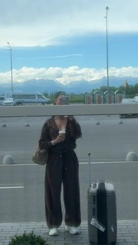 A woman stands in front of a reflective surface, holding a coffee cup and filming herself with a phone. She is wearing a brown tracksuit and a white top. A grey suitcase is on her right. Behind her, a parking lot, a white van, and mountains are visible under a partly cloudy sky.