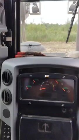 The video shows the interior of a bulldozer cab. The dashboard has gauges, including a speedometer and fuel gauge, and air vents. Outside the cab, a dirt construction site with vegetation is visible. The bulldozer appears to be parked or moving slowly on the site. There are no people visible inside the cab.