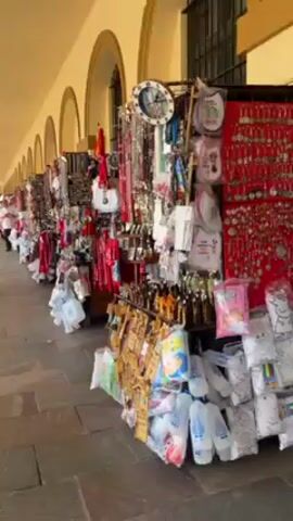 A sidewalk lined with vendor stalls is shown in the video. The stalls are filled with souvenirs, including jewelry, scarves, and baby items. The area has arched ceilings and is possibly a market or shopping area. A few people walk through the area, browsing the merchandise. The lighting is bright and sunny, suggesting it is daytime.
